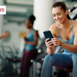 Frau im Sportoutfit sitzt im Fitnessstudio auf einem Trainingsgerät und lächelt beim Blick auf ihr Smartphone.