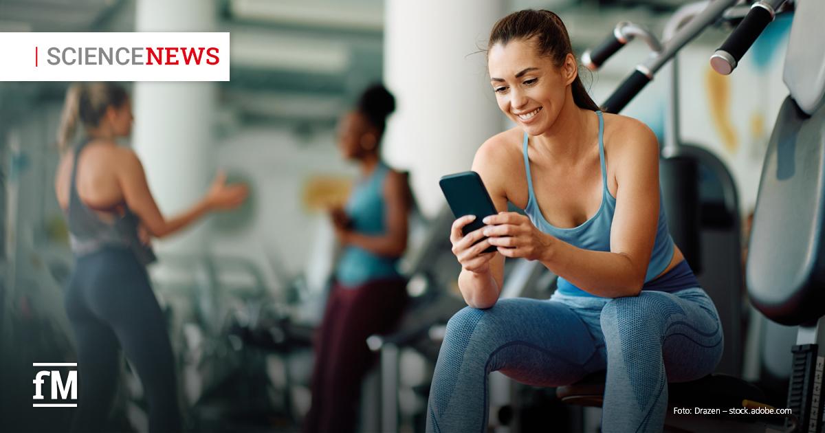 Frau im Sportoutfit sitzt im Fitnessstudio auf einem Trainingsgerät und lächelt beim Blick auf ihr Smartphone.