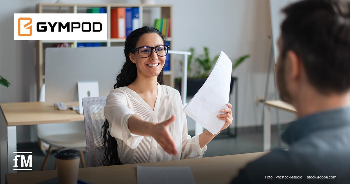 Eine Frau mit Brille sitzt an einem Schreibtisch in einem modernen Büro, reicht einem Mann mit dunklen Haaren, der im Anschnitt von hinten zu sehen ist, die Hand und hält in der anderen Hand Bewerbungsunterlagen. Sie lächelt und zeigt dabei die Zähne. Links oben befindet sich das GYMPOD-Logo auf einer weißen Fläche.
