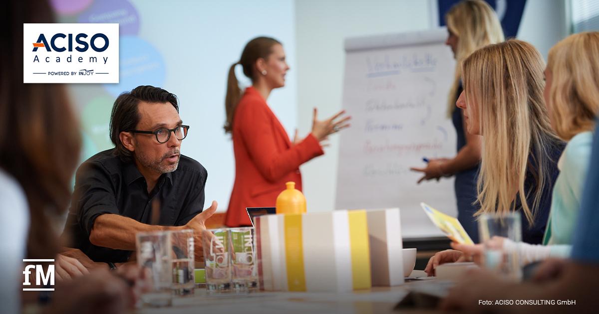 Menschen in einem Seminarraum bei einer Schulung der ACISO Academy; ein Mann spricht engagiert, während im Hintergrund eine Frau an einem Flipchart präsentiert.