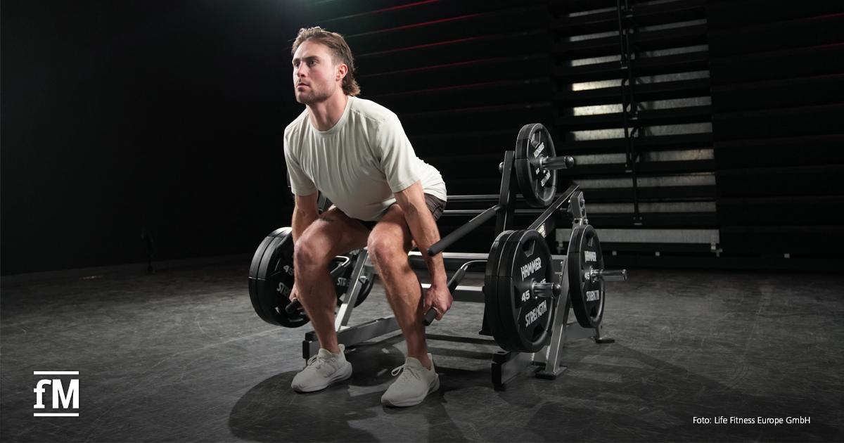 Ein junger Mann mit hellen Sportschuhen, Shorts und T-Shirt trainiert in einem dunklen Raum mit einer Hammer Strength-Maschine. Er ist in gehockter Position und hält das Gerät fest, als würde er eine Kreuzhebeübung ausführen.