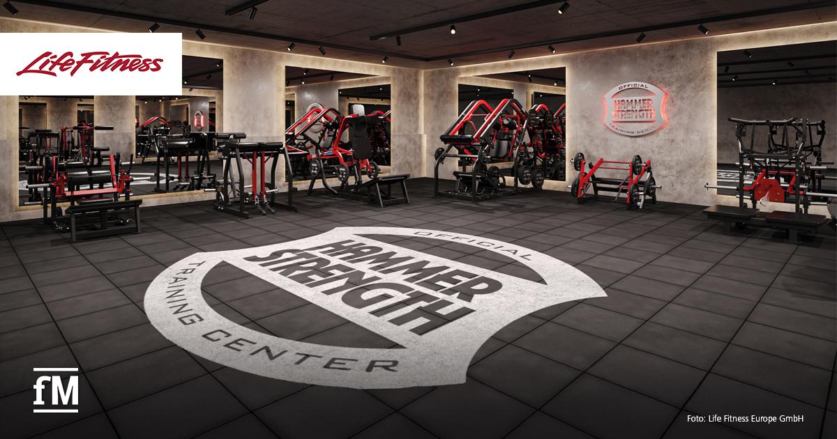 Moderner Trainingsraum mit zahlreichen roten und schwarzen Hammer Strength Geräten. In der Mitte des Raumes ist ein großer, runder Schriftzug 'Hammer Strength Training Center' auf dem Boden zu sehen.