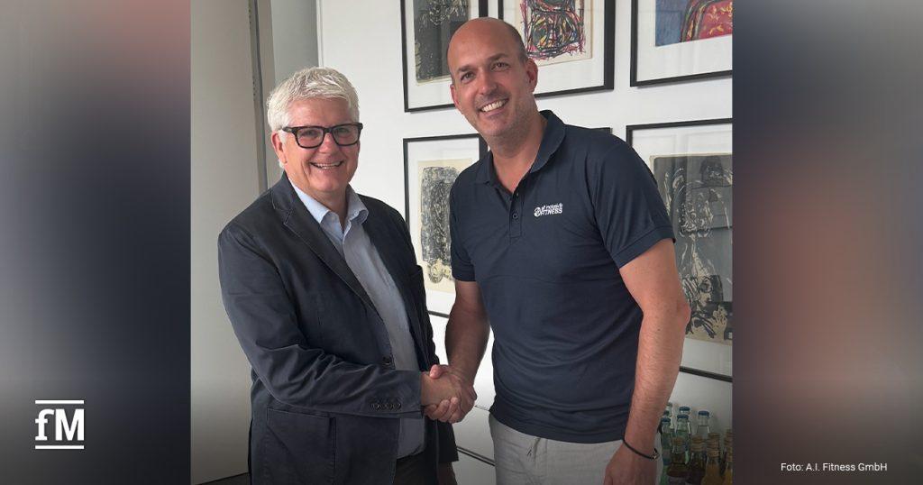 Zwei Männer stehen in einem Büro nebeneinander und geben sich die Hand. Der Mann links (Dr. Karsten Heppner) trägt eine Brille und ein dunkles Sakko, der Mann rechts (Stephan Schulan) trägt ein Polohemd mit Firmenlogo. Im Hintergrund hängen mehrere gerahmte Kunstwerke an der Wand.