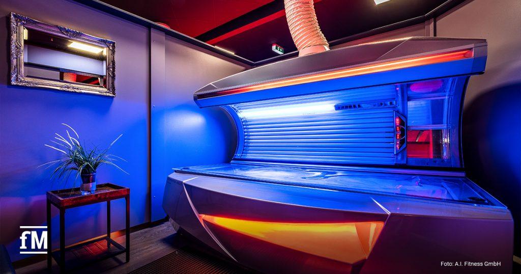 Ein Solarium steht in einem Raum mit blauen und orangefarbenen Lichtakzenten. Rechts davon befindet sich ein Lüftungsschlauch. Links davon steht ein kleiner Tisch mit einer Zimmerpflanze. An der Wand hängt ein Spiegel.