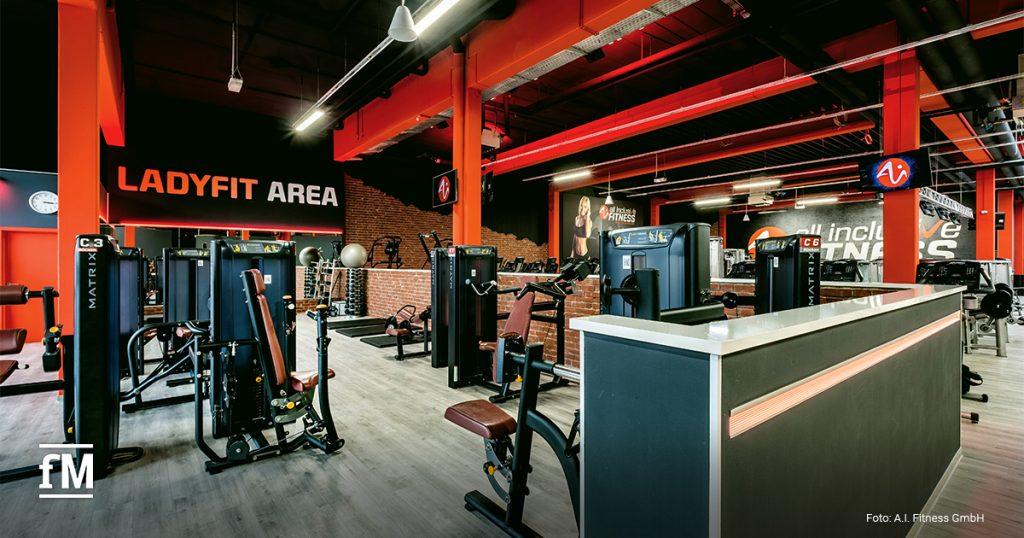 Ein Fitnessbereich mit mehreren Kraftgeräten steht in einem modern gestalteten Raum. Der Bereich ist mit der Aufschrift 'Ladyfit Area' versehen. Die Geräte sind schwarz-rot, Wände und Decke haben rote und schwarze Designelemente.