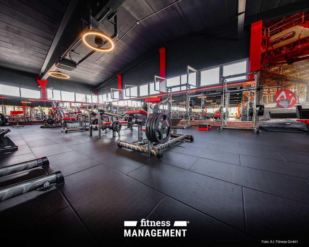 Krafttrainingsbereich mit Langhanteln, Gewichten und Squat-Racks auf schwarzem Gummiboden; Spiegelwand im Hintergrund, rote Akzente an Geräten und Säulen.