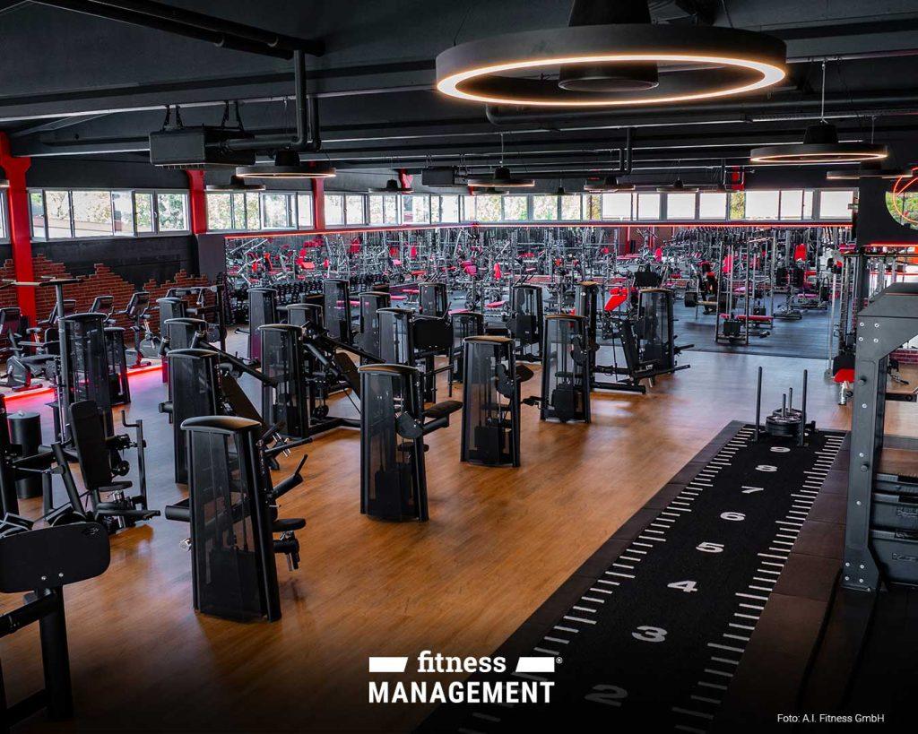Großer, moderner Fitnessbereich mit zahlreichen Kraftgeräten, dunklem Boden, roten Akzenten und beleuchtetem Rundlicht an der Decke; im Vordergrund ein Sprintstreifen mit Markierungen.