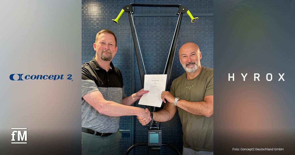 Zwei Männer (links Glenn Dwyer und Christian Toetzke) stehen in der Bildmitte vor einem Fitnessgerät (SkiErg), reichen sich die Hand und halten gemeinsam ein unterzeichnetes Dokument. Beide lächeln in die Kamera. Links ist das Logo von Concept2 und rechts das von HYROX vor unscharfem Hintergrund.