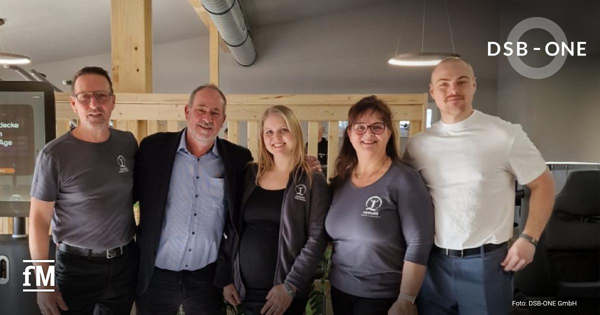 Fünf Personen stehen lächelnd nebeneinander in einem modern eingerichteten Therapieraum der Physiopraxis Hermes. Drei tragen Kleidung mit dem Logo 'Hermes Physio Point', eine Person trägt ein weißes T-Shirt, eine weitere Person (Jürgen Adams, 2. von links) trägt Anzugjacke.