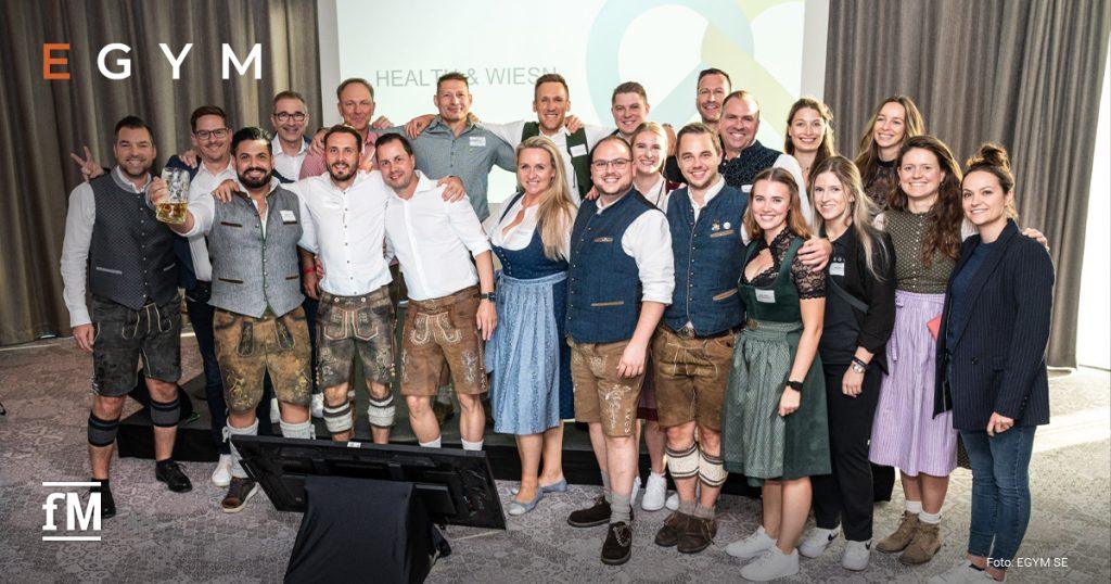 Eine große Gruppe von Personen in Trachtenkleidung steht vor einer Leinwand und lächelt in die Kamera.