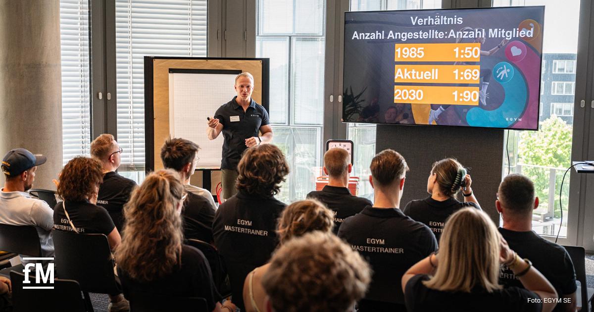 Ein Mann präsentiert vor einem Publikum eine Folie mit der Entwicklung des Betreuungsverhältnisses von Mitarbeitenden zu Mitgliedern, Zahlen und Jahresangaben sind auf einem großen Bildschirm zu sehen.