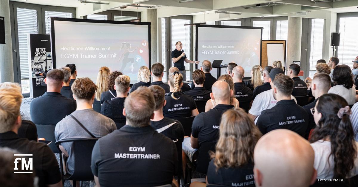 Ein Mann steht auf einer Bühne vor einer großen Leinwand und spricht zu einem vollbesetzten Raum mit Teilnehmer:innen, die alle schwarze EGYM-Shirts tragen.