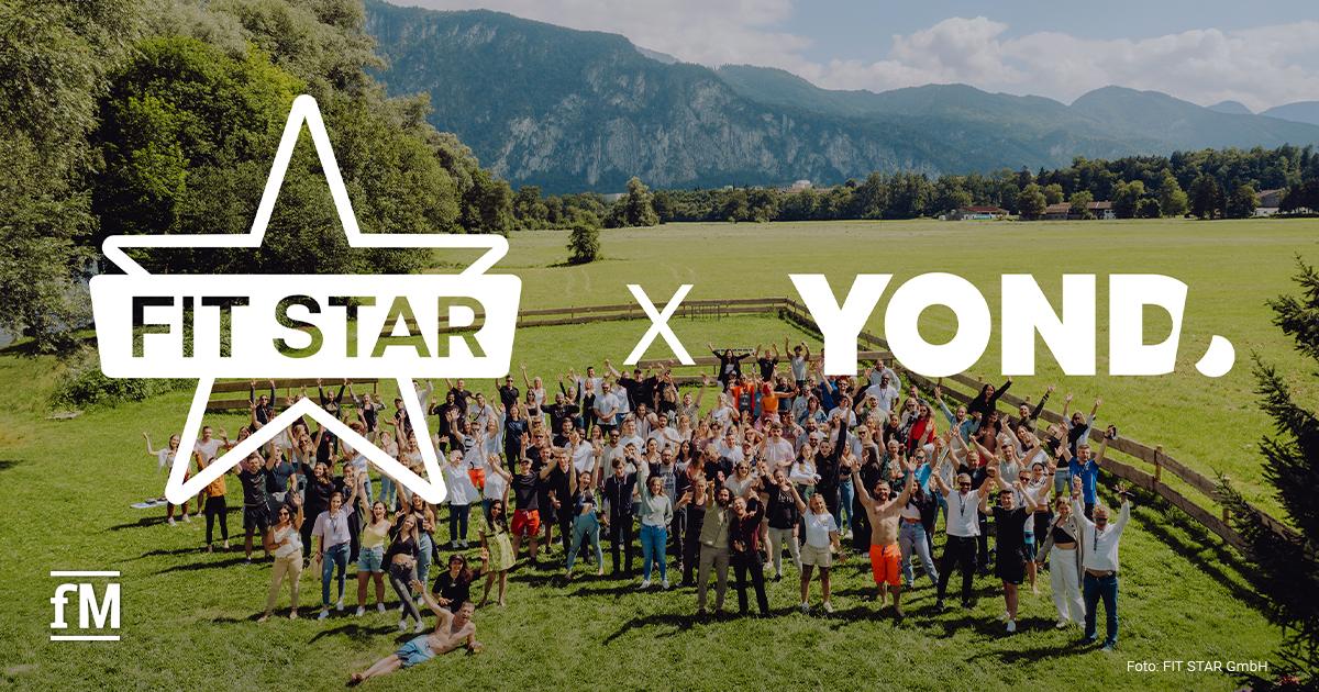 Eine große Gruppe von Menschen steht auf einer grünen Wiese mit erhobenen Armen, im Hintergrund Berge und Bäume. Im Vordergrund die Logos von FIT STAR und YOND.