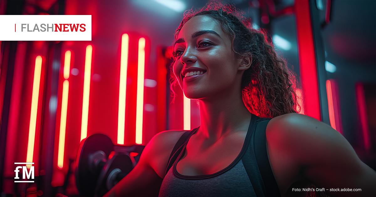 Eine junge Frau mit Sportkleidung trainiert an Hanteln in einem Fitnessstudio mit roter Beleuchtung. Sie lächelt und blickt nach vorne. Links unten befindet sich das fM-Logo und oben in der Ecke der Reiter 'Flashnews', der erste Teil grau, der zweite rot.