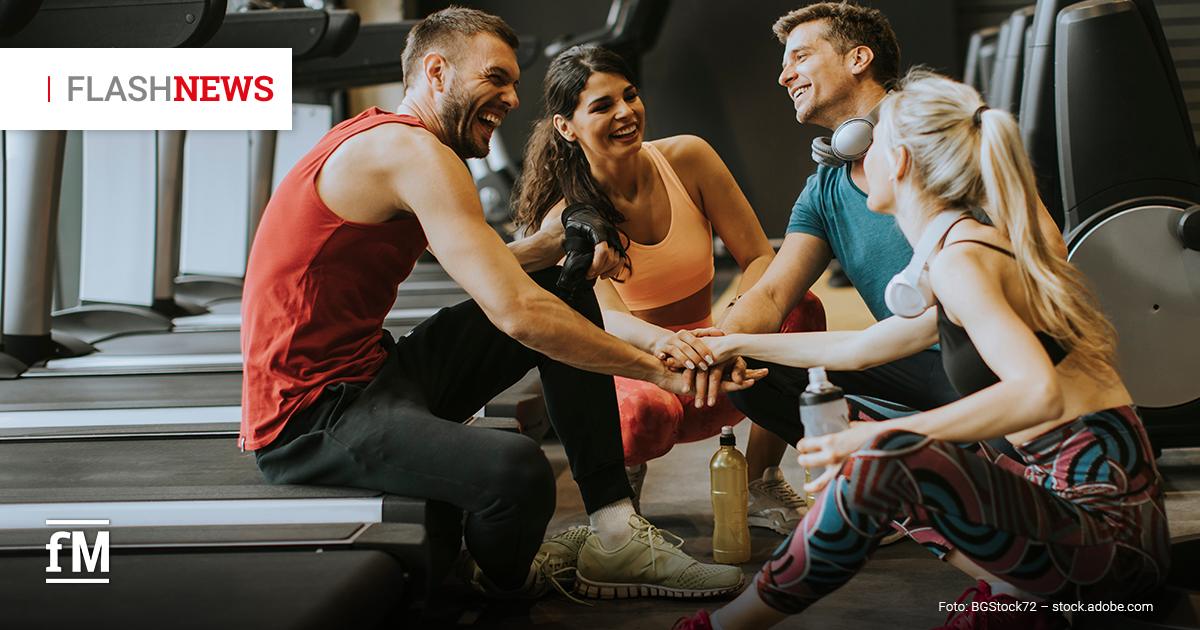 Vier junge Erwachsene in Sportkleidung sitzen lachend auf dem Boden eines Fitnessstudios zwischen Laufbändern und legen motiviert die Hände in der Mitte übereinander.