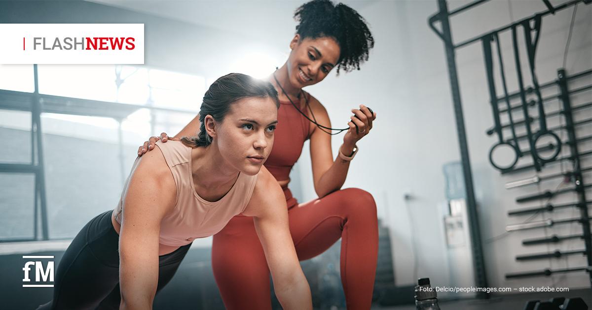 Zwei Frauen in Sportkleidung in einem Fitnessstudio: Eine macht auf einer Matte eine Plank-Übung, die andere kniet daneben, stoppt die Zeit mit einer Stoppuhr und legt unterstützend eine Hand auf ihre Schulter.