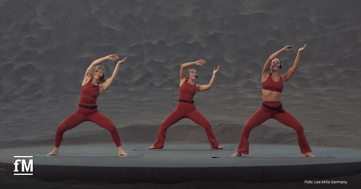 Drei Frauen in roten Sportoutfits stehen auf einer Bühne mit sandfarbenem Hintergrund, Arme seitlich erhoben in einer Tanzpose.