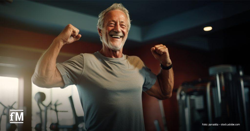 Älterer Mann mit grauem Haar, Bart und T-Shirt steht lachend im Fitnessstudio und zeigt seine Muskeln. Im Hintergrund stehen Fitnessgeräte.