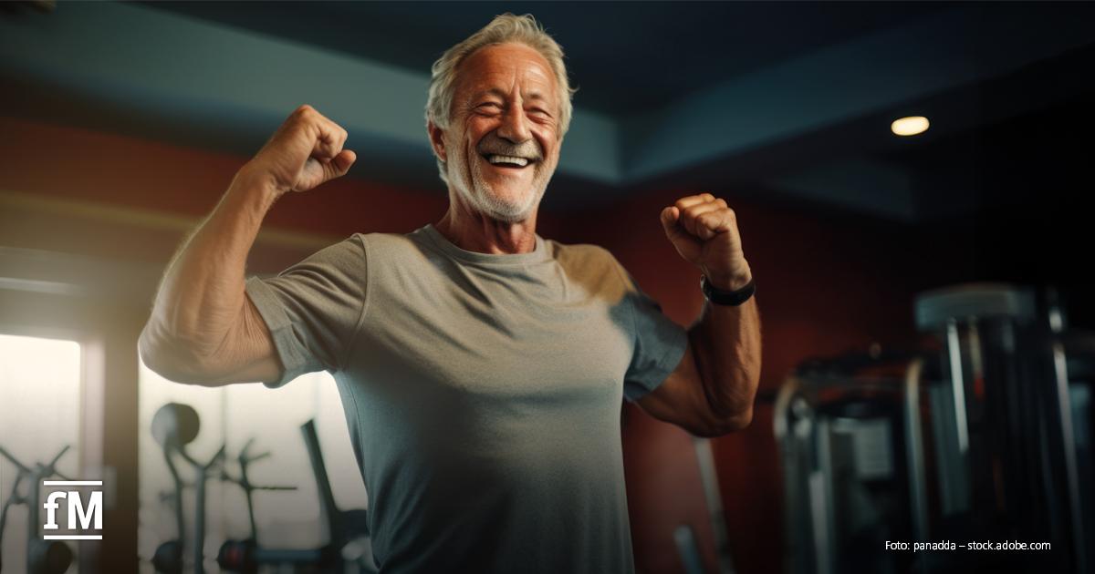 Älterer Mann mit grauem Haar, Bart und T-Shirt steht lachend im Fitnessstudio und zeigt seine Muskeln. Im Hintergrund stehen Fitnessgeräte.