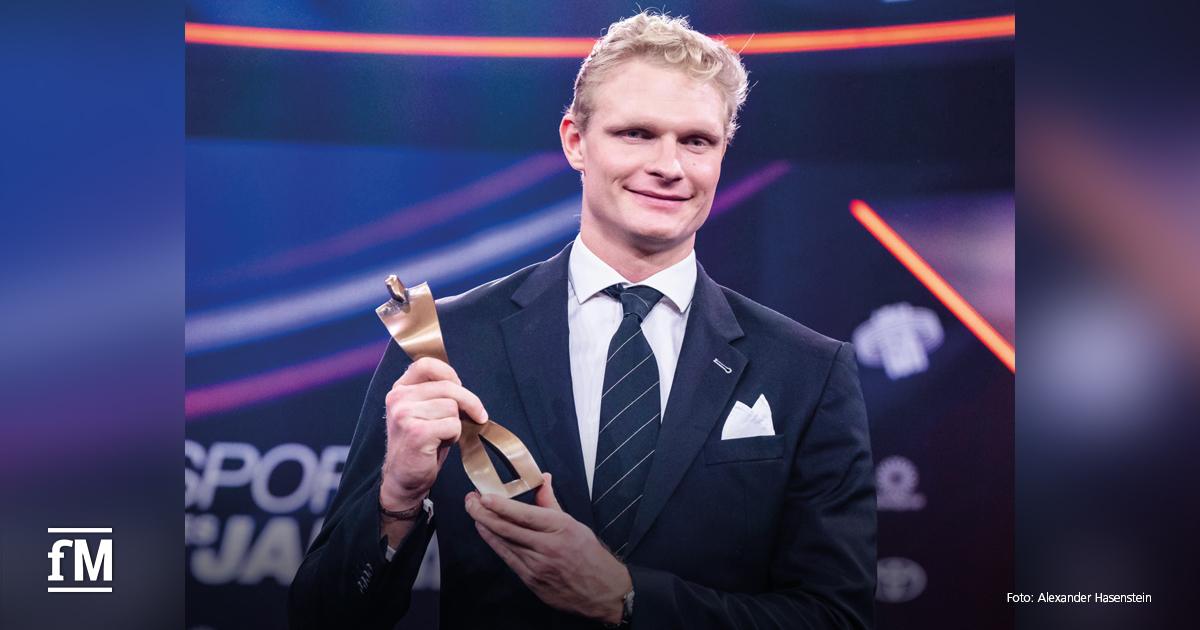 Ein blondhaariger Mann im Anzug steht auf einer Bühne und hält eine goldfarbene Trophäe in der Hand. Er lächelt, im Hintergrund sind Lichtinstallationen zu sehen.