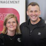Zwei Personen stehen nebeneinander und lächeln in die Kamera. Links eine Frau mit schulterlangen blonden Haaren, Brille und schwarzer Jacke mit dem Logo „fM – fitness MANAGEMENT“. Rechts ein Mann in schwarzer Adidas-Sportjacke mit weißen Streifen. Im Hintergrund sind ein rotes Schild mit weißer Schrift „WISSEN DAS DIE“ sowie ein graues Banner mit weißen Buchstaben zu sehen.