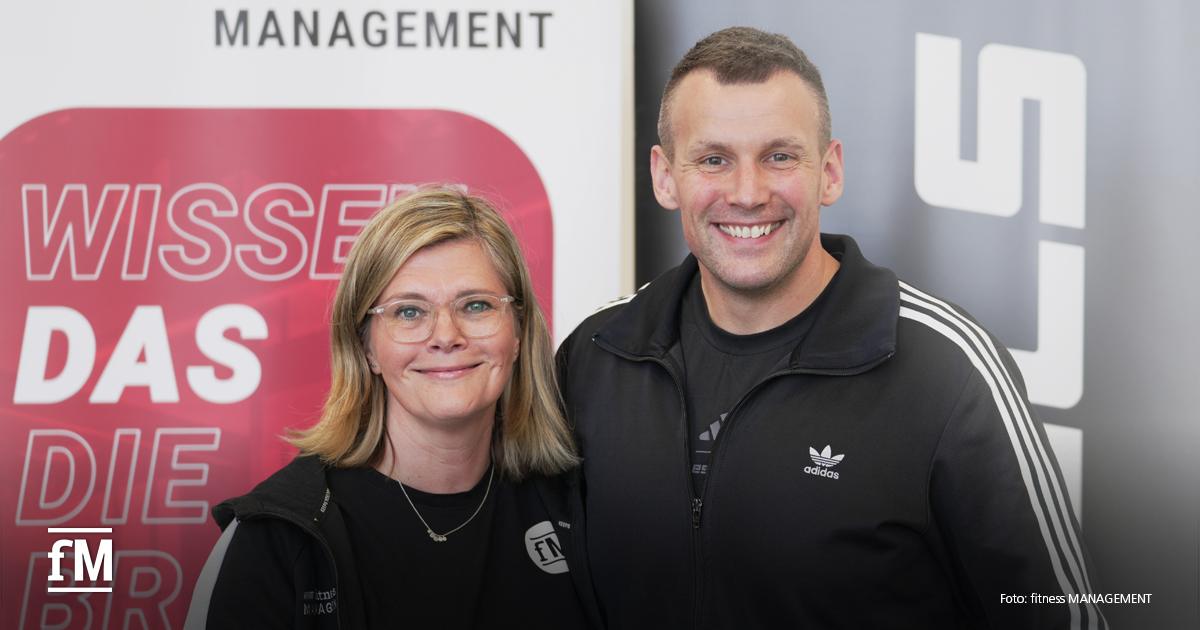 Zwei Personen stehen nebeneinander und lächeln in die Kamera. Links eine Frau mit schulterlangen blonden Haaren, Brille und schwarzer Jacke mit dem Logo „fM – fitness MANAGEMENT“. Rechts ein Mann in schwarzer Adidas-Sportjacke mit weißen Streifen. Im Hintergrund sind ein rotes Schild mit weißer Schrift „WISSEN DAS DIE“ sowie ein graues Banner mit weißen Buchstaben zu sehen.
