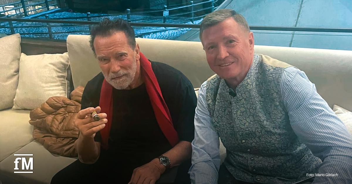 Arnold Schwarzenegger und Mario Görlach sitzen auf einer beigefarbenen Couch, Schwarzenegger, mit grauem Bart eine Zigarre in der Hand, trägt ein schwarzes Shirt mit rotem Schal. Görlach sitzt rechts neben ihm mit hellem Haar und einem bestickten Trachten-Gilet, beide blicken in die Kamera.
