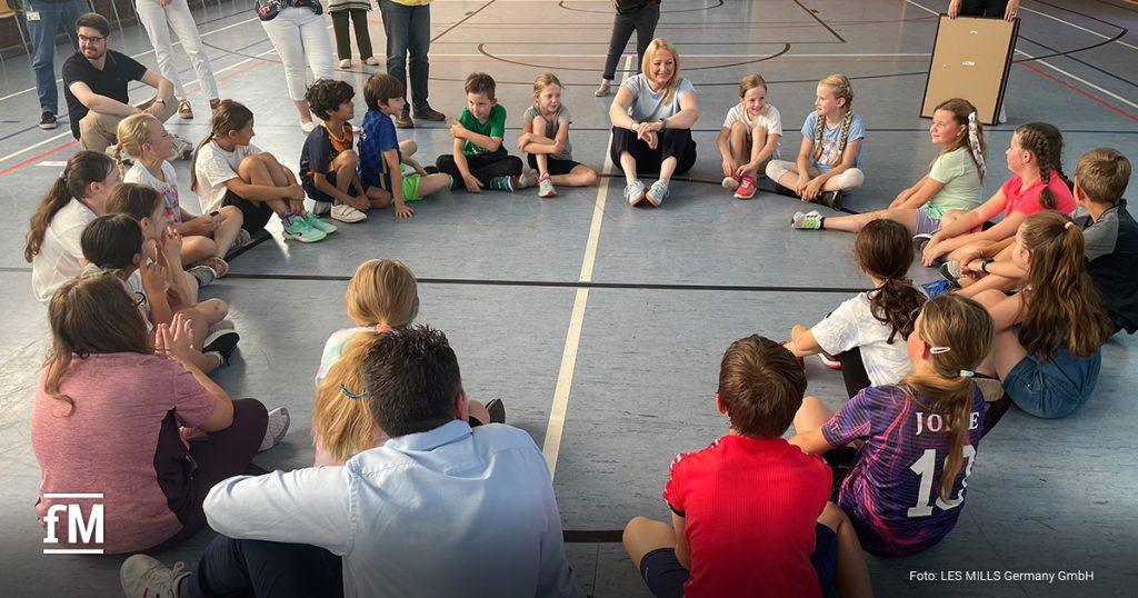 Kinder sitzen mit einer Lehrerin in einem großen Kreis in der Turnhalle, einige Erwachsene beobachten die Szene im Hintergrund, lockere und fröhliche Atmosphäre.