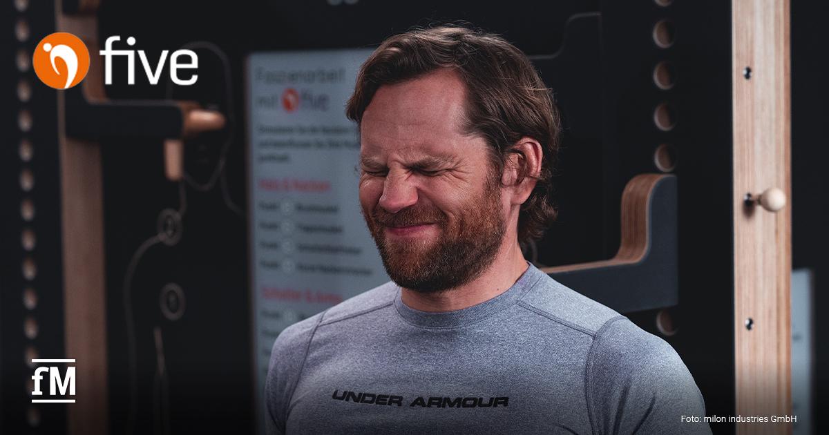 Mann mit grauem Funktionsshirt steht vor einer Trainingswand mit geschlossenen Augen und verzieht das Gesicht, als ob er eine intensive Dehnung oder Druck ausübt.