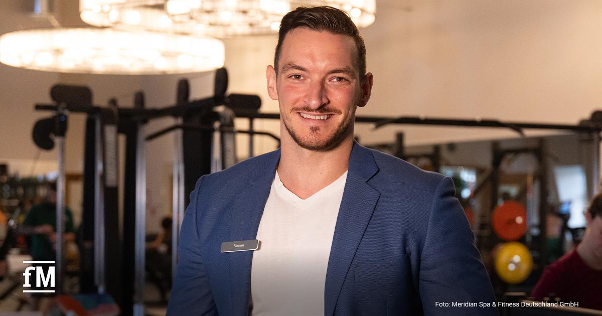Mann im blauen Sakko mit Namensschild 'Florian' steht lächelnd im Fitnessbereich mit Hanteln und Trainingsgeräten im Hintergrund.