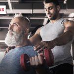 Ein älterer Mann mit grauem Vollbart trainiert in einem Fitnessstudio mit roten Kurzhanteln, während ein jüngerer Trainer in ärmellosem Shirt hinter ihm steht und ihn unterstützt.