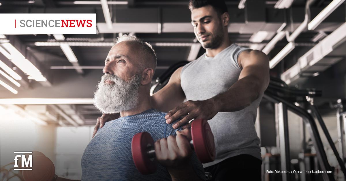 Ein älterer Mann mit grauem Vollbart trainiert in einem Fitnessstudio mit roten Kurzhanteln, während ein jüngerer Trainer in ärmellosem Shirt hinter ihm steht und ihn unterstützt.