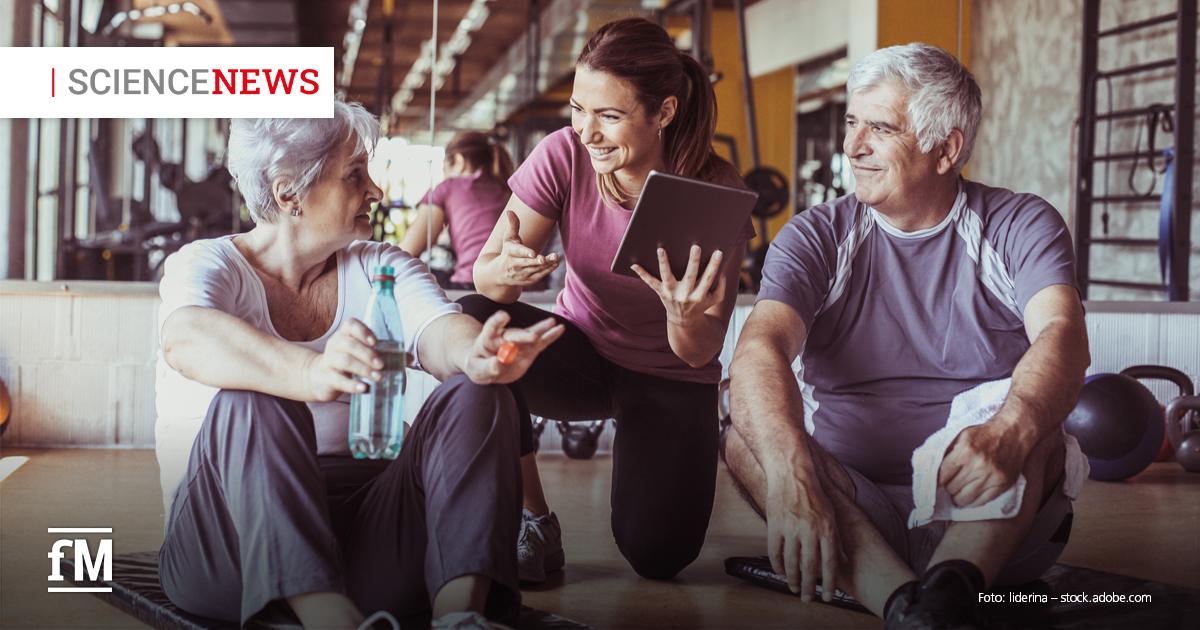 Zwei ältere Menschen in Sportkleidung sitzen in einem Fitnessstudio auf dem Boden und unterhalten sich lächelnd mit einer jüngeren Trainerin, die ein Tablet in der Hand hält.