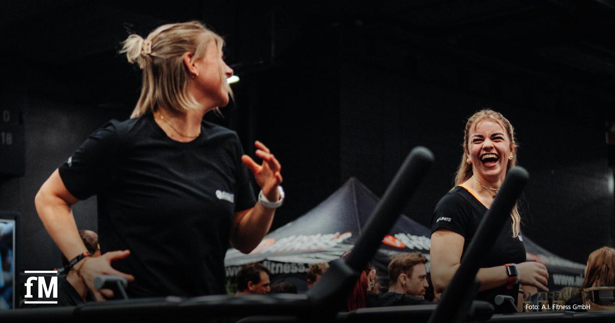 Zwei Frauen in schwarzen Sportshirts laufen nebeneinander auf Laufbändern und lachen, im Hintergrund Menschen und Messestände.