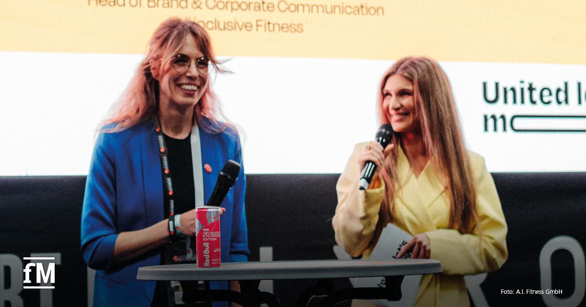 Zwei Frauen stehen auf einer Bühne, halten Mikrofone und lächeln. Eine trägt einen blauen Blazer, die andere einen gelben. Im Hintergrund Präsentationsfolie mit Text.