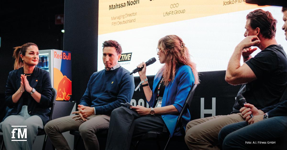 Vier Personen sitzen auf einer Bühne in einer Podiumsdiskussion. Eine Frau in blauem Blazer spricht ins Mikrofon, die anderen hören zu. Im Hintergrund Bildschirm mit Text.