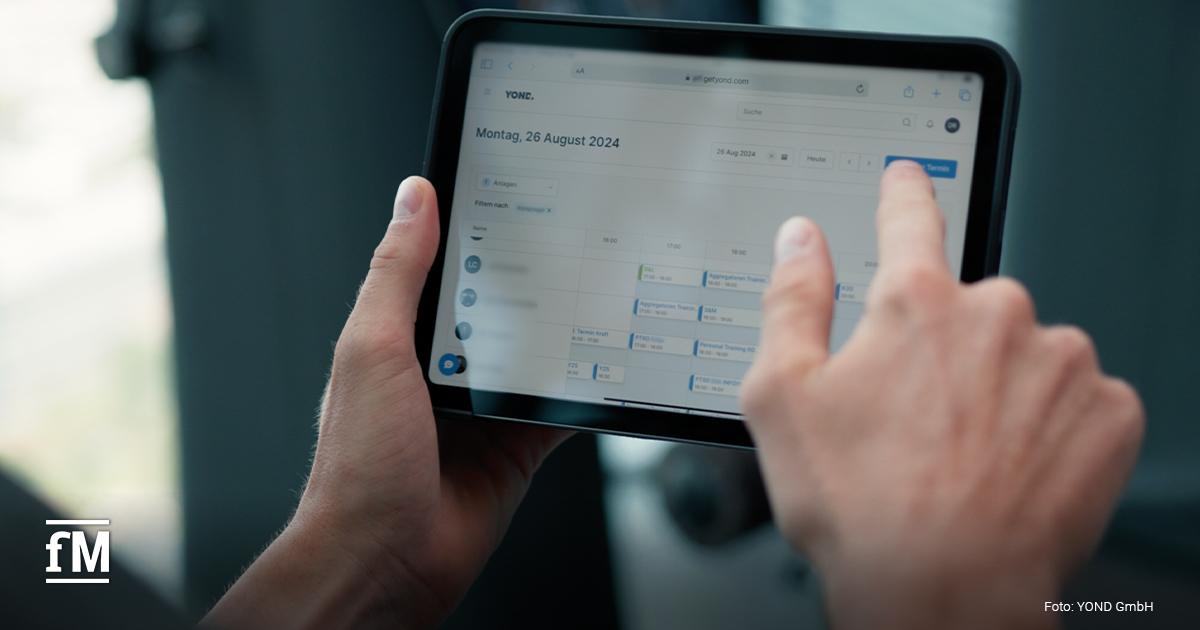 Nahaufnahme: Zwei Hände halten ein schwarzes Tablet und tippen auf den Bildschirm, der den YOND‑Kalender mit mehreren Terminkacheln für Montag, 26. August 2024 zeigt; im Hintergrund unscharf eine Glasfront.