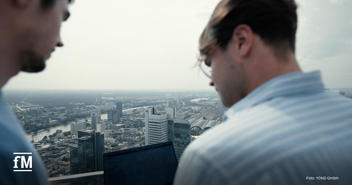 Blick von schräg hinten auf zwei Männer mit aufgeklapptem Laptop auf einer hohen Dachterrasse; im Hintergrund die Skyline von Frankfurt mit Main und Hochhäusern unter leicht bedecktem Himmel.