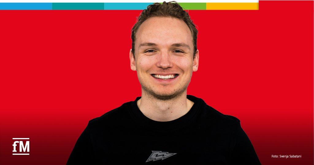 Porträt eines jungen Mannes, der freundlich in die Kamera lächelt. Er trägt ein schwarzes Oberteil vor einem roten Hintergrund mit buntem Farbstreifen oben.