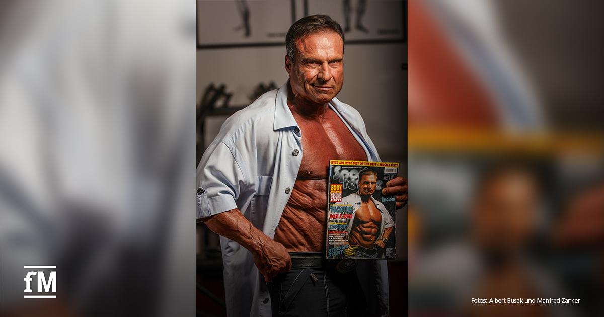 Ein muskulöser älterer Mann trägt ein offenes weißes Hemd und zeigt seine definierte Brust. In der Hand hält er eine Zeitschrift mit seinem eigenen Titelbild, das ihn in identischer Pose zeigt.
