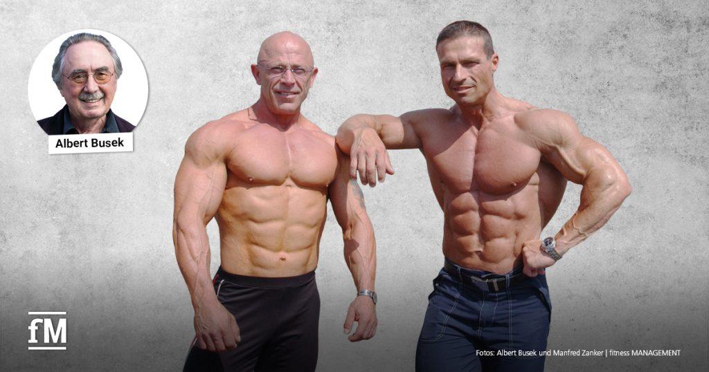 Zwei muskulöse Männer mit nacktem Oberkörper posieren nebeneinander vor grauem Hintergrund; einer stützt den Arm auf der Schulter des anderen.