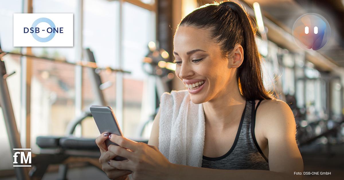 Junge Frau mit Handtuch über der Schulter sitzt im Fitnessstudio und lächelt beim Blick auf ihr Smartphone, während sie den digitalen Service MYCOACH von DSB-ONE nutzt.