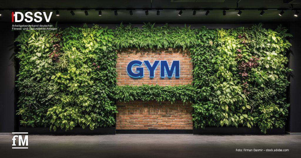 Leuchtendes blaues GYM-Schild auf roter Backsteinwand, umrahmt von dichter grüner Pflanzenwand.