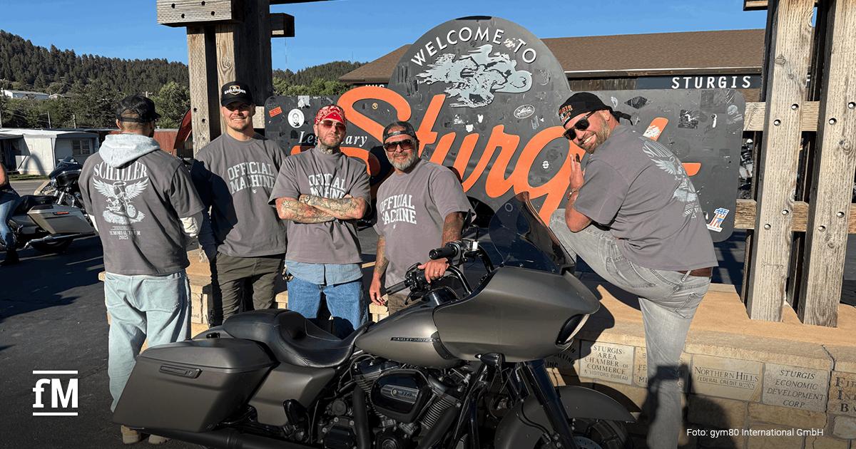 Fünf Männer posieren fröhlich vor dem 'Welcome to Sturgis'-Schild in South Dakota; alle tragen Shirts mit der Aufschrift 'Rainer Schaller Memorial Tour 2025', davor steht ein schwarzes Motorrad.