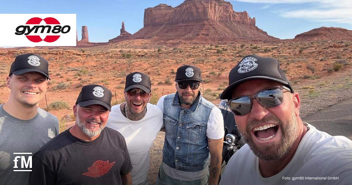 Fünf lachende Männer mit schwarzen Basecaps posieren bei sonnigem Wetter vor der roten Felsenlandschaft des Monument Valley im Südwesten der USA. Alle tragen Freizeitkleidung; im Hintergrund sind markante Tafelberge und das gym80-Logo zu sehen.