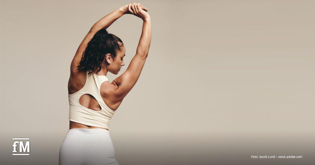 Eine Frau in Sportkleidung steht seitlich zum Betrachter und streckt ihre Arme über den Kopf, während sie sich zur Seite dehnt. Sie trägt ein cremefarbenes Sport-Top mit Rückenausschnitt und weiße Leggings.