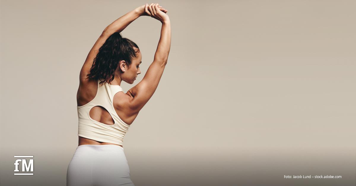 Eine Frau in Sportkleidung steht seitlich zum Betrachter und streckt ihre Arme über den Kopf, während sie sich zur Seite dehnt. Sie trägt ein cremefarbenes Sport-Top mit Rückenausschnitt und weiße Leggings.