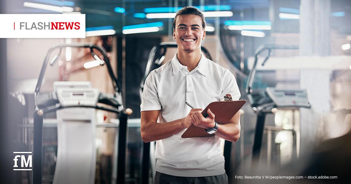 Ein junger Mann in weißem Polohemd in einem Fitnessstudio mit Klemmbrett und Stift; im Hintergrund Laufbänder und Fitnessgeräte.