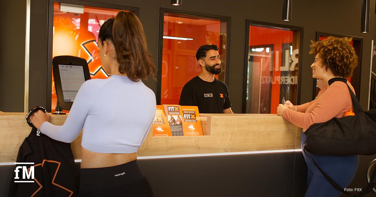 Zwei Frauen stehen an der Rezeption des Fitnessstudios FitX, eine checkt sich an einem Tablet ein, die andere spricht lächelnd mit einem Mitarbeiter hinter dem Tresen. Im Hintergrund steht in großen weißen Buchstaben „FOR ALL OF US“.