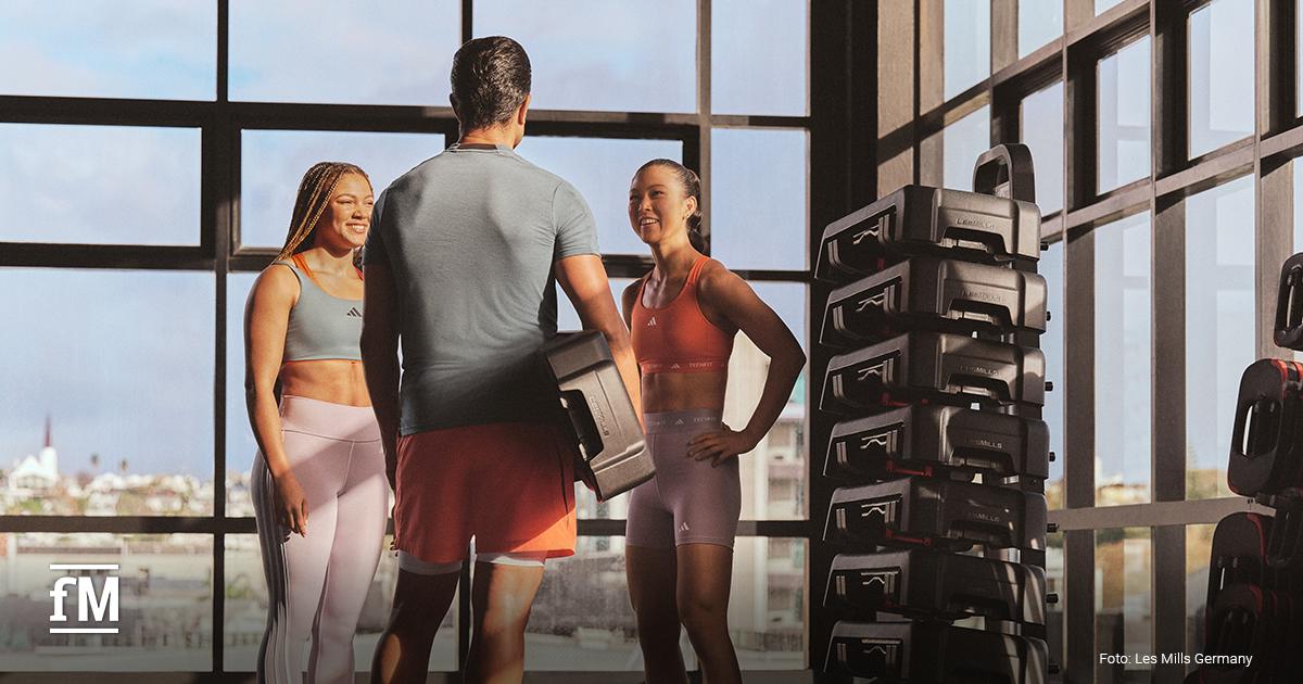 Drei Personen im Fitnessstudio stehen zusammen und sprechen, einer hält Trainings-Equipment.